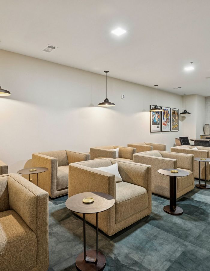 Modern lounge with beige armchairs, small tables, pendant lights, and a workspace at the back.