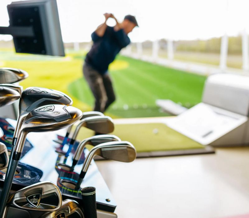 A set of golf clubs in focus with a person swinging at a driving range in the blurred background.