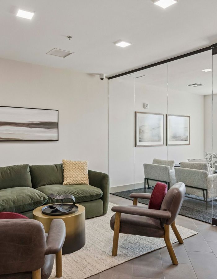 Modern lounge with green sofa, two chairs, a round table, and glass wall overlooking a meeting area.
