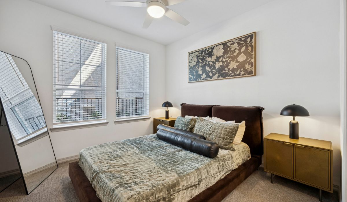 Modern bedroom with a velvet bed, gold nightstands, a large mirror, and two windows letting in natural light.