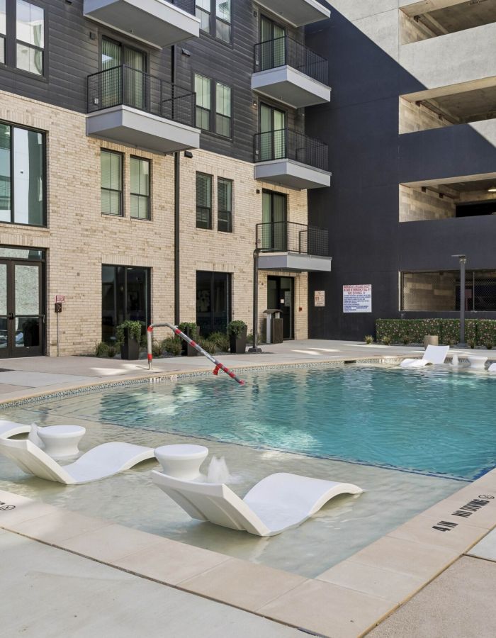Modern apartment pool area with lounge chairs in shallow water, surrounded by buildings and a parking garage.