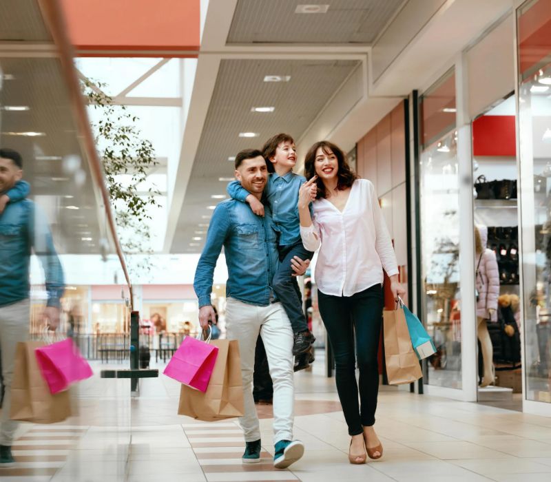 A smiling family with shopping bags walks through a mall; a child rides on the father's shoulders.