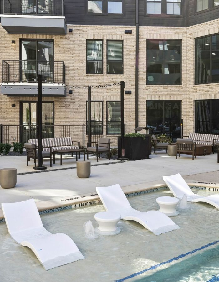 Modern apartment courtyard with pool, lounge chairs in water, outdoor seating, and string lights overhead.