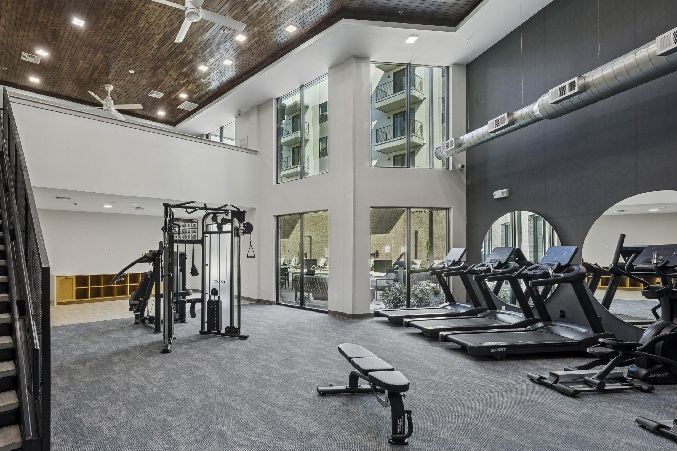 Modern gym with treadmills, weight machines, a bench, large windows, and high ceilings with ceiling fans.