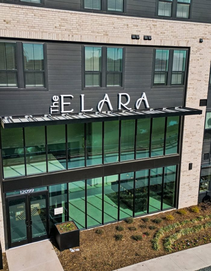 Modern apartment building entrance with large windows and a sign that reads "The Elara" above the doorway.