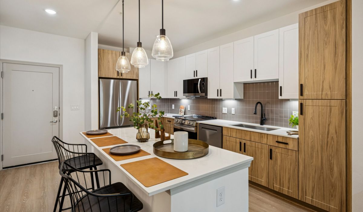 Modern kitchen with wood cabinets, white countertops, pendant lights, and two black barstools at an island.