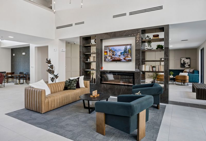 Modern lounge with tan sofa, blue chairs, a fireplace, built-in shelves, and a TV in a bright, open space.