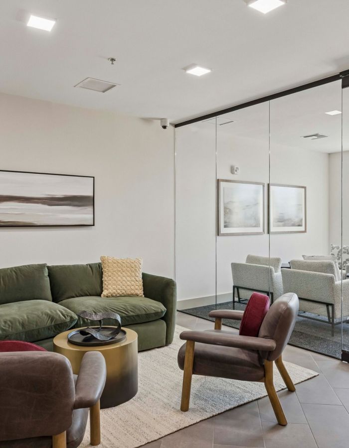 Modern lounge with green sofa, two chairs, a round table, and glass wall overlooking a meeting area.